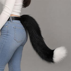 May include: A long, plush, faux fur animal tail accessory in solid black with a contrasting white tip. The tail is attached to a black belt worn around the waist, featuring thick, soft synthetic fibers that extend outward in a curved shape.
