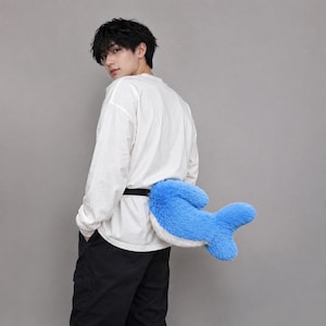 May include: A plush, bright blue and white whale tail accessory attached to a black adjustable strap. The tail features a soft, fuzzy texture with a rounded, bifurcated fin shape, designed to be worn around the waist as a decorative costume piece.