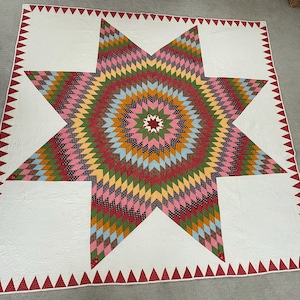 May include: A large square quilt featuring a central eight-pointed star composed of small diamond-shaped fabric patches in shades of pink, green, yellow, blue, and brown. The quilt has a white background and a border of red triangular shapes.