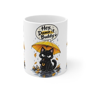 May include: A white ceramic mug featuring a graphic of a black cat holding a yellow umbrella in the rain. The cat sits in a puddle with falling leaves. Above the cat, a speech bubble contains the black and yellow text: Hey, Damn it Buddy.