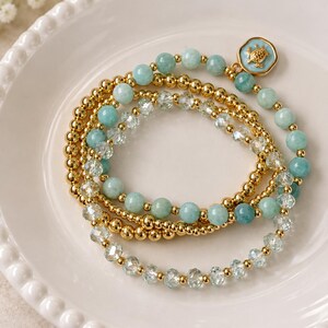 May include: A set of three stacked bracelets featuring polished gold-tone beads, light blue gemstone beads, and clear faceted crystal beads. One bracelet includes a small, round, light blue charm with a gold-tone sea turtle emblem on it.