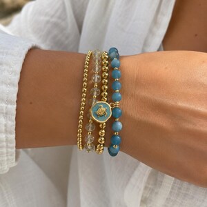 May include: A set of four stacked bracelets featuring a strand of small gold-tone beads, a strand of clear faceted beads, a strand of gold-tone beads with a central light blue circular charm depicting a gold sea turtle, and a strand of blue beads.