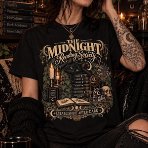 Könnte beinhalten: Ein schwarzes T-Shirt mit einem Grafikdruck, der den Text "THE MIDNIGHT Reading Society" und "ESTABLISHED AFTER DARK" zeigt. Das Design umfasst eine schwarze Katze, einen Bücherstapel, eine Kerze, einen Totenkopf, Kristalle und Efeuranken in Gold- und Grüntönen.