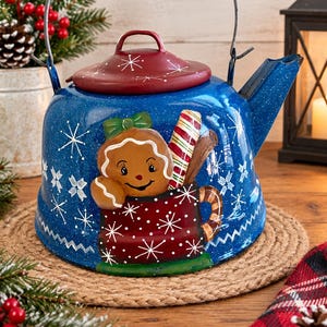 May include: A blue metal tea kettle with a speckled finish, featuring a red lid and a painted design of a gingerbread person in a red mug with a candy cane. The kettle is decorated with white snowflake patterns and has a metal handle.