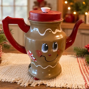 May include: A brown teapot designed to resemble a gingerbread person with a red lid, red handle, and red spout. The teapot features white icing-style trim, a painted face with large eyes and a smile, and a small peppermint candy graphic on the side.