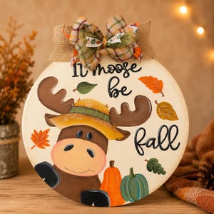 May include: A round wooden sign featuring a painted brown moose wearing a yellow hat, surrounded by orange and green pumpkins and autumn leaves. The text reads It moose be fall in black script. A plaid and burlap bow is attached to the top.
