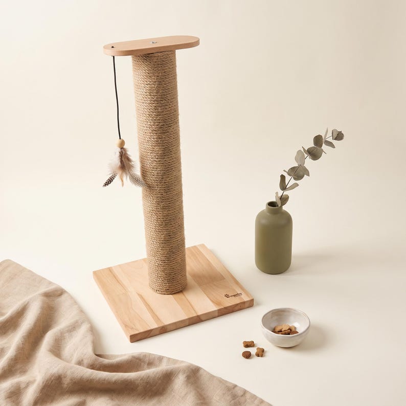 May include: A cat scratching post featuring a vertical sisal rope-wrapped column attached to a square light wood base. A dangling toy with feathers hangs from the top wooden platform. Nearby sits a matte olive green vase and a small ceramic bowl.