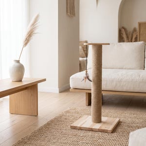 May include: A cat scratching post featuring a square light-colored wooden base and a vertical cylindrical column wrapped in natural tan sisal rope. A thin black string hangs from the top platform, attached to a small wooden bead and feathers.