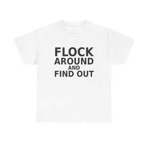 Puede incluir: Una camiseta blanca de manga corta con el texto negro FLOCK AROUND AND FIND OUT impreso en una fuente sans-serif en negrita en el centro del pecho. La prenda tiene un cuello redondo clásico y una silueta estándar de corte recto.