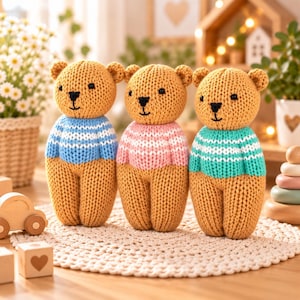 May include: Three hand-knitted teddy bears in tan yarn, each wearing a different colored sweater. The left bear wears a blue and white striped sweater, the middle bear a pink and white striped sweater, and the right bear a teal and white striped sweater.