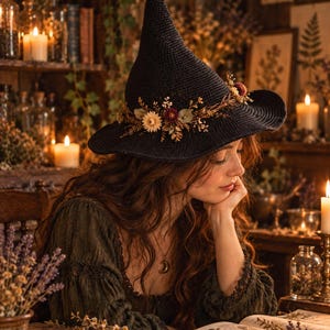 Puede incluir: Un sombrero negro de punta con ala ancha, que presenta una banda decorativa de flores secas y follaje en tonos crema, burdeos y verde. El sombrero tiene una apariencia tejida y texturizada, adornado con pequeños y delicados elementos botánicos.