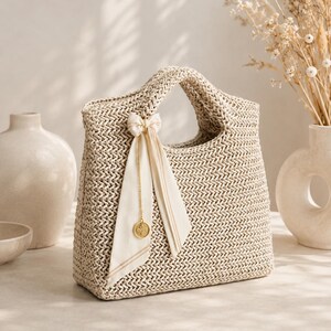 Op de afbeelding: Een beige gevlochten handtas met een gestructureerd silhouet en geïntegreerde handvatten. De tas is versierd met een crèmekleurig stoffen lint dat tot een strik is geknoopt, geaccentueerd door een dunne goudkleurige ketting en een kleine ronde metalen bedel.