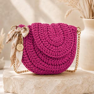 Puede incluir: Un bolso bandolera de ganchillo magenta con solapa redondeada, que cuenta con una correa de cadena de metal dorado y un pañuelo decorativo de aspecto sedoso en color beige atado a un lado. Un pequeño dije dorado con un trébol cuelga de la cadena.