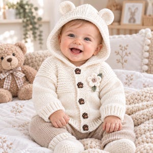 Pode incluir: Um casaco de malha cor de creme com capuz apresentando duas pequenas orelhas de urso. O casaco tem cinco botões castanhos em forma de pegadas e uma aplicação de flor de croché branca no peito, usado sobre calças de malha bege.