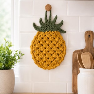 May include: A pineapple-shaped crochet potholder featuring a textured golden-yellow body with a raised stitch pattern and a dark green leafy top. The item includes a small loop at the top for hanging, showcasing a handcrafted design.
