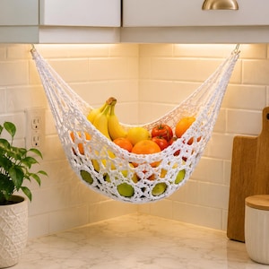 Puede incluir: Una hamaca de frutas tejida en crochet blanco suspendida bajo armarios de cocina, que contiene una variedad de productos frescos incluyendo plátanos amarillos, naranjas y tomates rojos, con un diseño de red de malla ancha y ganchos.
