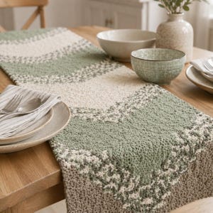 Puede incluir: Un camino de mesa texturizado con un patrón de chevrón repetitivo en tonos verde salvia, crema y beige. El camino está elaborado con un punto de tejido o ganchillo visible, creando una superficie gruesa y táctil sobre la mesa.