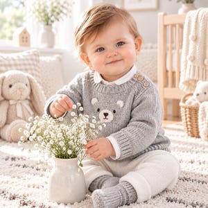 Könnte beinhalten: Ein hellgrauer Strickpullover mit einem Bärengesicht-Motiv auf der Brust und zwei kleinen Holzknöpfen an der Schulter. Der Pullover wird über einem weißen Hemd mit Kragen getragen, kombiniert mit einer weißen Strickleggings und grauen Socken.