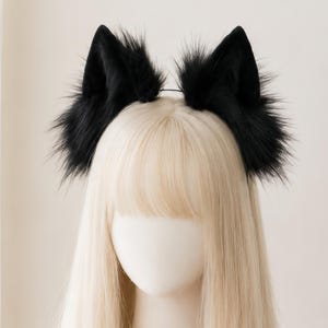 Puede incluir: Una diadema con dos orejas de gato puntiagudas y erguidas, cubiertas de pelo sintético negro, largo y denso. Las orejas están unidas a una banda fina de color oscuro que descansa sobre una cabeza con cabello rubio claro liso y flequillo.