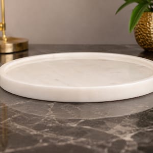 May include: A circular white marble tray with a smooth, polished surface and a raised rim. The tray features subtle grey veining throughout the stone, creating a natural pattern across the flat base and the thick, rounded edge of the object.