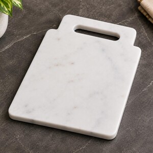 May include: A rectangular white marble serving board with subtle grey veining and a smooth, polished finish. The board features a flat surface and an integrated handle cutout at the top center, providing a functional grip for easy transport.