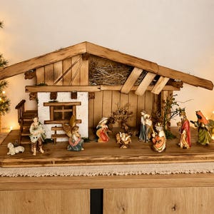 Könnte beinhalten: Eine hölzerne Weihnachtskrippe mit einem rustikalen Stall, einer weißen Wand und einem Strohdach. Das Set enthält handbemalte Figuren von Maria, Josef, dem Jesuskind in der Krippe, einem Engel, Hirten und den drei heiligen Königen auf einem Sockel.