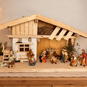 Könnte beinhalten: Eine hölzerne Weihnachtskrippe mit einem rustikalen Stallgebäude inklusive Heuboden, einem weiß verputzten Hausbereich und handbemalten Figuren, darunter die heilige Familie, ein Engel, die drei Könige und verschiedene Tiere.