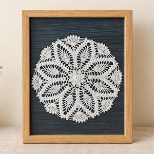 Pode incluir: Um centro de mesa de crochê branco de formato circular com um intrincado padrão floral e de abacaxi, exibido dentro de uma moldura de madeira castanho-clara sobre um fundo azul escuro texturizado. O centro apresenta um motivo central.