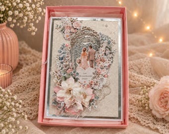 Peut inclure: Une carte de vœux rectangulaire dans une boîte rose présentant un design 3D superposé avec un couple sous une arche. La carte est ornée de décorations florales roses et blanches, d'accents argentés et d'une étiquette en forme de cœur avec le texte Love is the most beautiful journey.