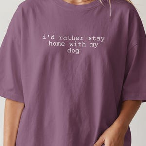 Peut inclure: Un t-shirt à manches courtes de couleur prune avec le texte blanc "i'd rather stay home with my dog" imprimé dans une police de style machine à écrire sur la poitrine. Le vêtement présente un col rond et une coupe ample et décontractée.
