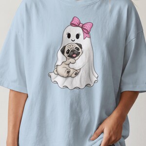 May include: A light blue t-shirt featuring a graphic of a white ghost wearing a pink polka dot bow. The ghost is holding a tan pug puppy with a black face. The illustration is centred on the chest of the shirt, showcasing a playful design.