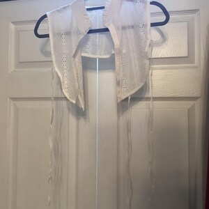 May include: A white detachable collar made of sheer fabric with intricate eyelet embroidery and scalloped edges. The collar features a pointed design with a front button closure and long, thin white fabric ties extending from the sides.