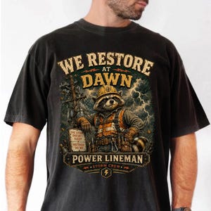 Puede incluir: Una camiseta negra con el gráfico de un mapache vestido como técnico de líneas eléctricas con casco y chaleco de seguridad. El texto dice WE RESTORE AT DAWN, POWER LINEMAN, STORM CREW y THE STORM DOESN'T SLEEP NEITHER DO WE sobre un fondo de tormenta.