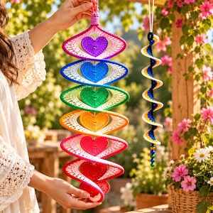 May include: A vertical hanging decoration featuring a series of six crocheted hearts in rainbow colors, each framed by a white spiral structure. Beside it hangs a smaller, slender spiral ornament in blue, yellow, and white.