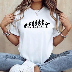 Puede incluir: Una camiseta blanca de manga corta con un estampado gráfico negro que representa la evolución de la humanidad, comenzando desde un primate y progresando a través de varias etapas hasta una figura final que realiza un pino de breakdance.