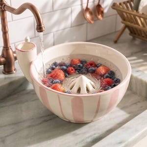 Puede incluir: Un cuenco colador de cerámica con rayas rosas verticales y un exprimidor central, lleno de fresas y arándanos frescos que se enjuagan bajo un grifo de cobre. El cuenco cuenta con un asa lateral y un interior blanco con orificios.