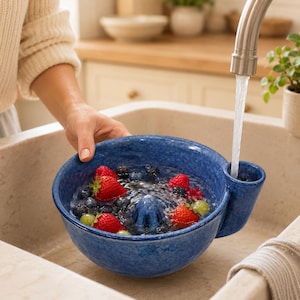 Könnte beinhalten: Eine blaue Beeren-Seiher-Schüssel aus Keramik mit gesprenkelter Oberfläche, einem zentralen Abflusskegel und einem seitlichen Ausguss. Die Schüssel ist mit Wasser gefüllt, in der frische rote Erdbeeren und Blaubeeren unter einem Wasserhahn gespült werden.