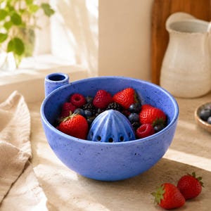 May include: A blue ceramic citrus juicer bowl with a speckled finish, featuring a central reamer and a small handle. The bowl is filled with fresh red strawberries, blackberries, and blueberries, with two additional strawberries resting nearby.