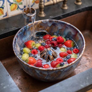 Peut inclure: Une passoire à baies en céramique émaillée ronde avec une finition mouchetée bleue et brune, dotée d'un cône de drainage central et d'une poignée latérale. Le bol est rempli d'eau, de fraises fraîches, de framboises, de myrtilles et de raisins verts.