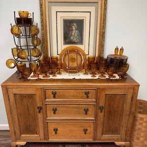 May include: A collection of amber-colored pressed glass tableware including various sizes of goblets, plates, and cups arranged on a wooden sideboard. A metal rack holds stacked cups, while a framed portrait sits in the center behind the glassware.
