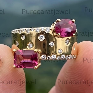 May include: A wide gold band ring featuring a scattered pattern of small round white diamonds, a large oval-cut pink gemstone, and a rectangular emerald-cut pink gemstone. The ring has a polished gold finish and a textured, multi-stone design.