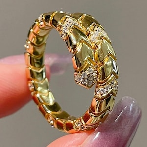 May include: A gold ring shaped like a coiled snake with a textured, scale-like pattern along the band. The head and sections of the body are encrusted with small, clear, sparkling stones that catch the light, creating a detailed, metallic finish.