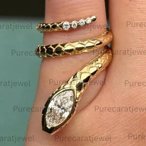 May include: A gold snake-shaped wrap ring featuring a textured scale pattern along the band. The head of the snake is set with a single marquise-cut white gemstone, while the tail end is accented with four small round-cut white gemstones.