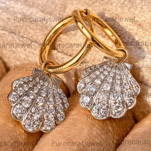 May include: A pair of gold hoop earrings featuring dangling seashell-shaped charms. Each shell charm is encrusted with numerous small, clear, sparkling stones that cover the entire surface, creating a bright, reflective, and textured appearance.