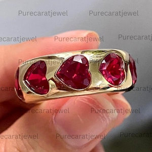 May include: A gold band ring featuring a series of vibrant red heart-shaped gemstones set flush into the metal surface. The ring displays a smooth, polished finish with multiple heart-shaped red stones arranged horizontally around the band.