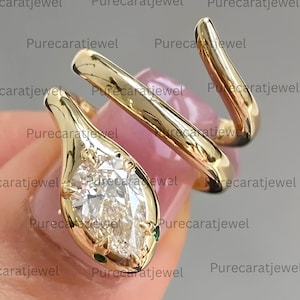 May include: A gold bypass ring shaped like a snake, featuring a marquise-cut clear gemstone set in the head and two small green stones for eyes. The polished metal band wraps around the finger in a spiral design with the text Purecaratjewel.