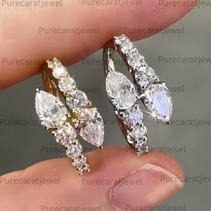 May include: Two hoop earrings featuring a bypass design with pear-cut and round-cut clear stones. One earring is set in yellow metal and the other in white metal. The text Purecaratjewel is repeated across the image. The stones are faceted and reflective.