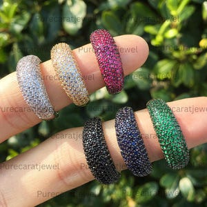 May include: Six dome-shaped rings featuring pavé-set stones in white, yellow, pink, black, dark blue, and green. Each band is densely covered in small, round, faceted gemstones that create a uniform, sparkling texture across the entire surface.