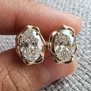 May include: A pair of gold stud earrings featuring oval-cut clear stones set in a scalloped, organic-shaped metal frame. The earrings have a polished finish and a reflective surface, showcasing a faceted design that catches the light.