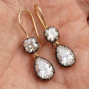 May include: A pair of drop earrings featuring two clear faceted stones each, set in dark metal bezels with gold-toned wire hooks. The upper stone is square-shaped, while the lower stone is pear-shaped, creating a dangling design.